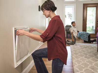 Woman replacing air filter in HVAC system, demonstrating routine maintenance for improved indoor air quality and system efficiency, with child playing in the background.
